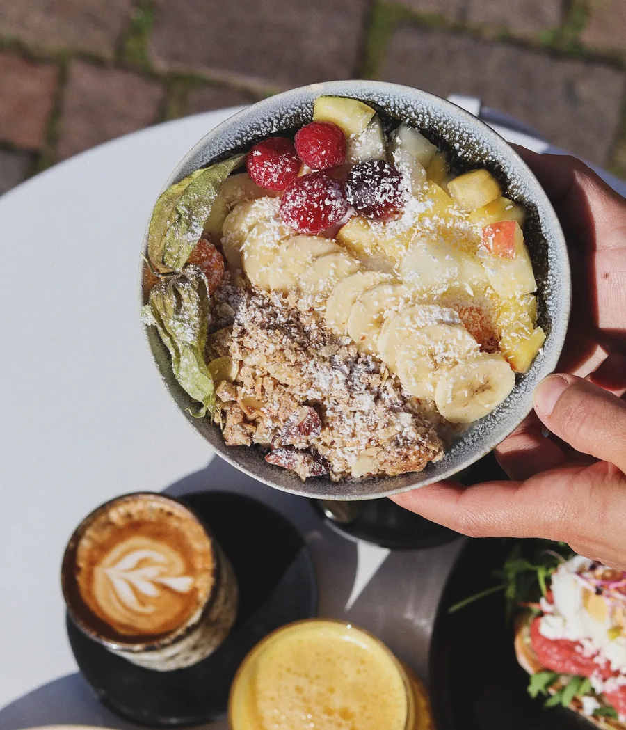 Bowl mit Granola, Banane, Beeren und Kokosraspeln, dazu Cappuccino und Saft