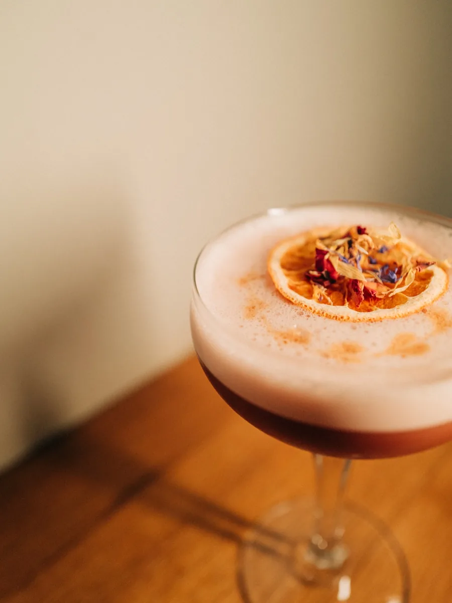 Sour im Coupe-Glas mit Schaumkrone, getrockneter Orangenscheibe und Blüten auf Holztisch