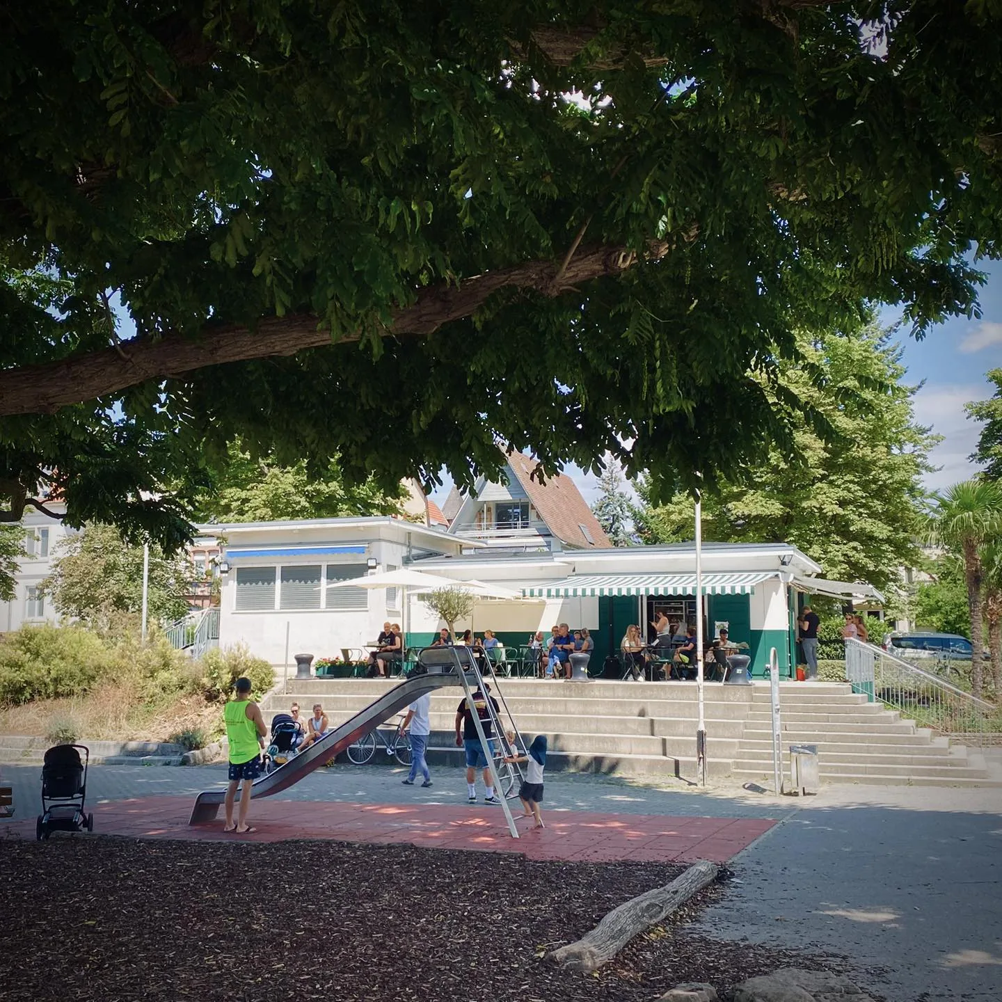 Der Kiosk do Bairro mit grün-weißer Markise unter einem großen Baum, davor der Wasserspielplatz mit Rutsche und Familien an den Tischen