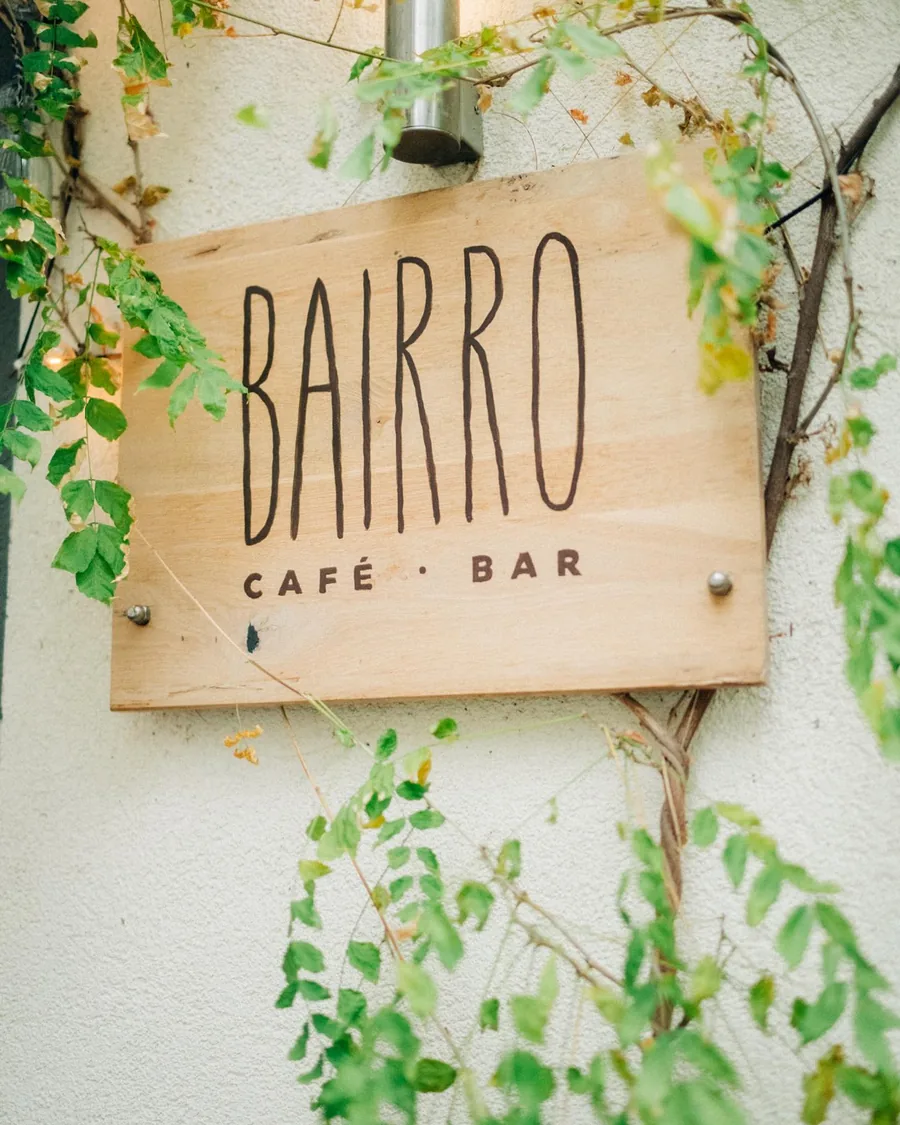 Das handgezeichnete Holzschild „BAIRRO Café · Bar“ an der Hauswand, umrankt von Efeu