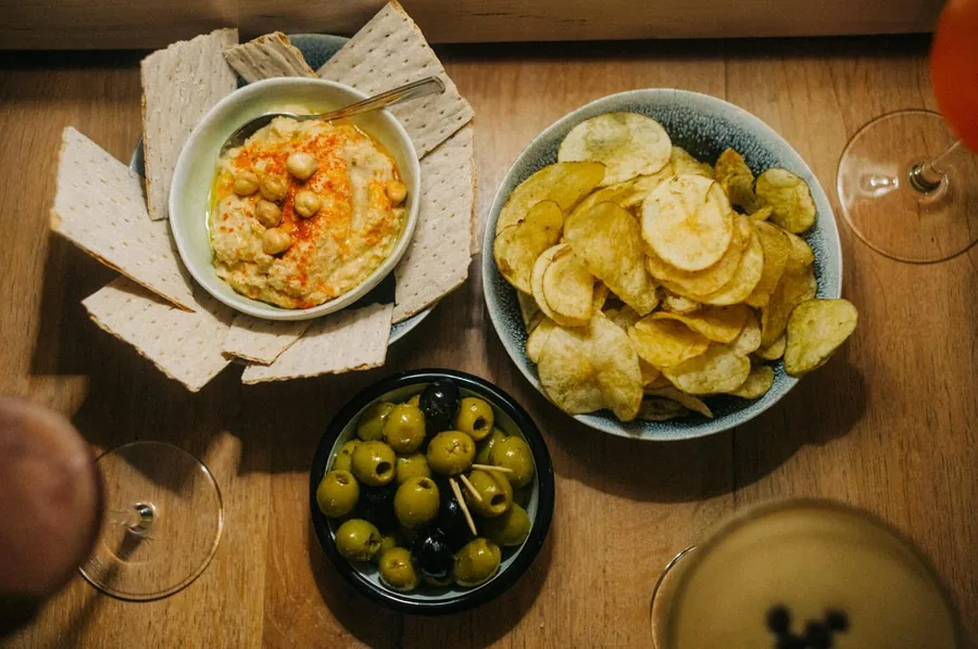 Hummus, Oliven und gesalzene Kartoffelchips auf einem Holztisch, daneben zwei Gläser